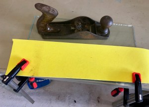 120 grit sandpaper clamped to table and glass ready to clean the sole of the plane